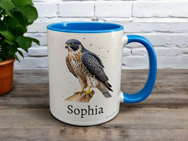 Blue & White Ceramic Mug with Falcon Design sat on a table. The Cup has a standard handle. Customisable text is printed below the Falcon design in a black text.