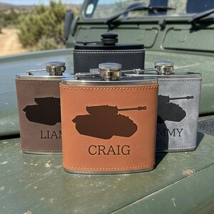 May include: Four stainless steel flasks wrapped in faux leather in brown, tan, grey, and black. Each flask features a dark silhouette of a tank and a personalised name engraved on the front. The metal caps are silver or black with a screw-top design.