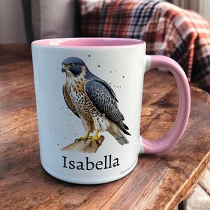 Pink & White Ceramic Mug with Falcon Design sat on a table. The Cup has a standard handle. Customisable text is printed below the Falcon design in a black text.