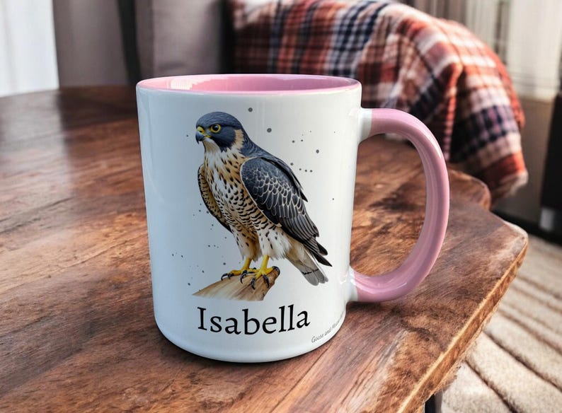 Pink & White Ceramic Mug with Falcon Design sat on a table. The Cup has a standard handle. Customisable text is printed below the Falcon design in a black text.