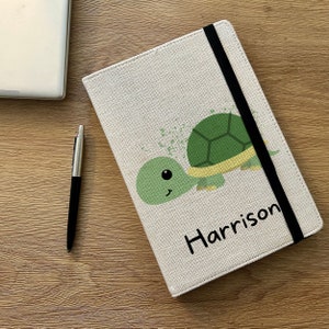 May include: A beige linen notebook with a black elastic closure. The cover features a cartoon turtle with green and yellow accents and the name "Harrison" printed in black.