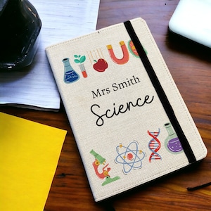 May include: A white notebook cover with a black elastic closure. The cover features colourful science-themed illustrations, including a beaker, a microscope, an atom, and a DNA strand. The text "Mrs. Smith Science" is printed in black on the cover.