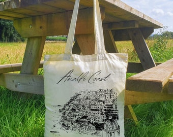 May include: A white canvas tote bag with black text that reads "Amalfi Coast". The bag features a black and white illustration of the Amalfi Coast.