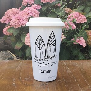 Może przedstawiać: Biały kubek podr&oacute;żny z czarno-białym rysunkiem dw&oacute;ch desek surfingowych i fal. Kubek ma nadrukowane imię "James" z przodu.