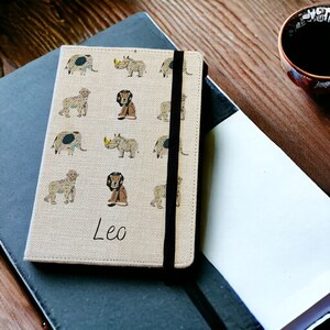May include: A beige linen notebook with a black elastic closure. The cover features a repeating pattern of illustrated animals, including lions, elephants, and rhinos. The name "Leo" is written in cursive on the front of the notebook.
