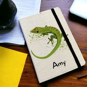 May include: A beige canvas notebook with a green gecko illustration and the name "Amy" printed on the front. The notebook has a black elastic closure.