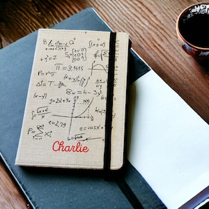 May include: A beige notebook with a black elastic closure. The cover is printed with mathematical equations and a graph. The name "Charlie" is written in red at the bottom of the notebook.
