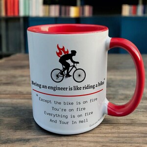 May include: White ceramic mug with a red interior and handle. The mug features a black graphic of a cyclist with flames above the head, and text that reads "Being an engineer is like riding a bike..." and other text.