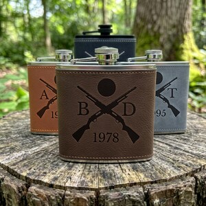 May include: Four metal flasks wrapped in leather-like material in brown, tan, grey, and black. Each flask features a laser-engraved design of two crossed shotguns, a circular icon, two letters, and the year 1978, 1995, or other dates.