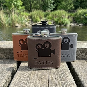 May include: Four metal flasks wrapped in leather-like material in brown, tan, grey, and black. Each flask features a laser-engraved vintage movie camera icon and a personalised name, including the names Darren, Timo, and Isaac, with a screw-top lid.