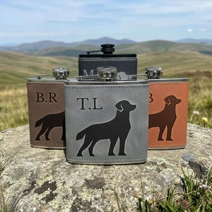 May include: Four metal flasks wrapped in leather-like material in shades of brown, grey, and black. Each flask features a silhouette of a dog and engraved initials, including B.R, T.L, and M. The flasks have a screw-top lid and a rectangular shape.