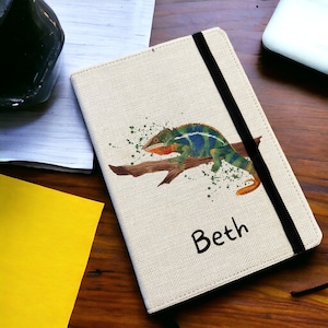 May include: A beige linen notebook cover with a black elastic closure. The cover features a colourful chameleon illustration with green, blue, and orange accents. The name "Beth" is printed in black on the cover.