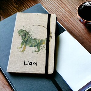 May include: A beige linen notebook with a black elastic closure. The cover features a watercolour illustration of a green iguana with a splatter effect. The name "Liam" is printed in black below the iguana.