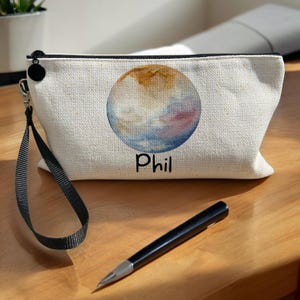 May include: A white canvas zippered pouch with a black zipper and a black strap. The pouch features a watercolour painting of a planet in shades of brown, yellow, blue, and pink. The text "Phil" is printed in black below the planet.