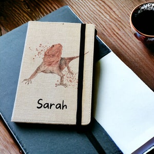 May include: A beige linen notebook with a black elastic closure. The cover features a watercolour illustration of a red-bearded dragon with brown spots. The name "Sarah" is printed in black below the illustration.