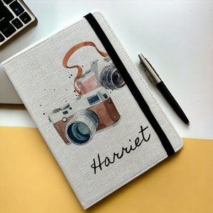 May include: A white canvas notebook with a black elastic closure and a watercolour illustration of vintage cameras. The notebook is personalised with the name "Harriet".
