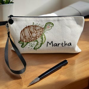 May include: A white canvas zippered pouch with a black zipper and a black strap. The pouch features a watercolour illustration of a green sea turtle with a brown shell and the name "Martha" printed in black.