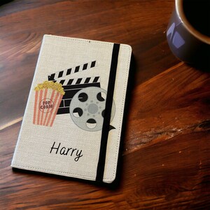 May include: A white linen notebook with a black elastic closure. The cover features a black and white illustration of a film reel, clapperboard, and a bucket of popcorn. The name "Harry" is printed in black below the illustration.