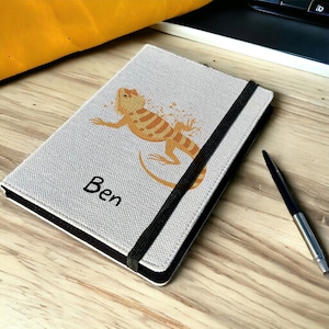 May include: A light grey notebook featuring an orange and yellow lizard illustration. The name "Ben" is printed in black. A black elastic band holds the notebook closed. A pen lies on the wooden surface next to the notebook.