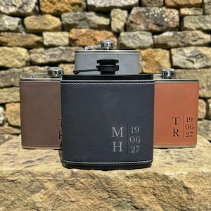 May include: Four rectangular metal flasks wrapped in faux leather in shades of dark grey, brown, tan, and light grey. Each flask features a screw-top lid and is engraved with initials and the date 19 06 27, with white stitching along the bottom edge.