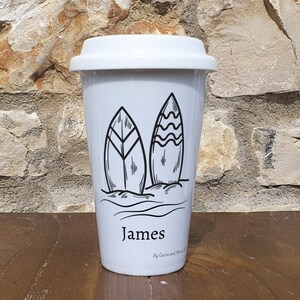 Może przedstawiać: Biały ceramiczny kubek podr&oacute;żny z czarnym konturem dw&oacute;ch desek surfingowych i fal. Kubek ma nadrukowane imię "James" z przodu i tekst "By Goose and Moose" nadrukowany pod imieniem.