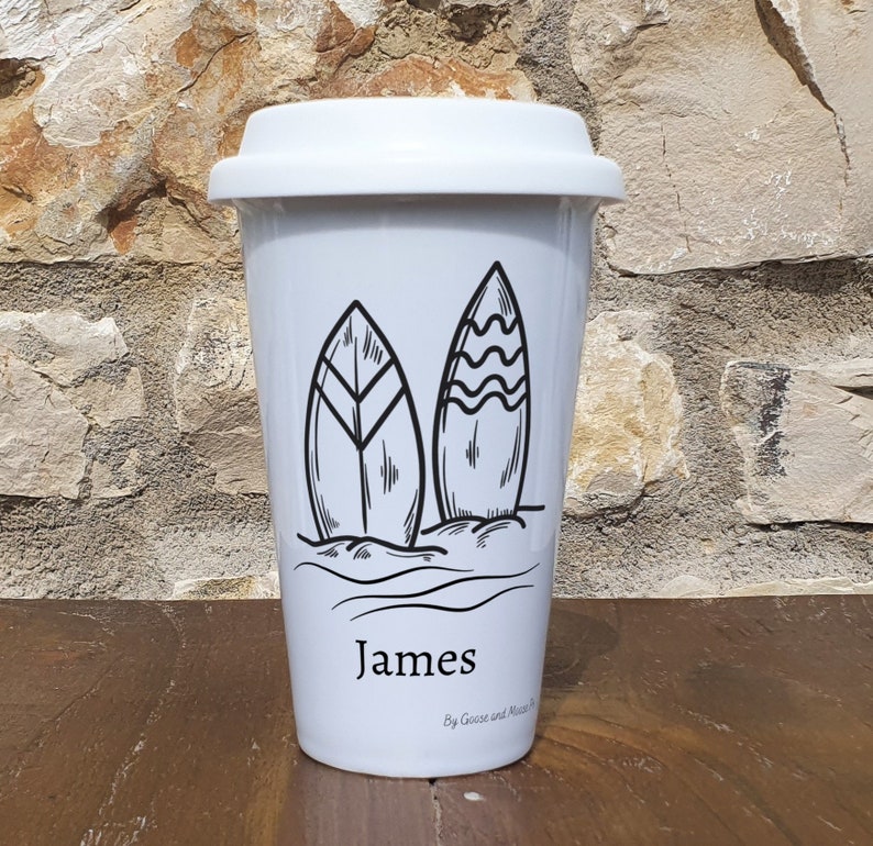 Może przedstawiać: Biały ceramiczny kubek podr&oacute;żny z czarnym konturem dw&oacute;ch desek surfingowych i fal. Kubek ma nadrukowane imię "James" z przodu i tekst "By Goose and Moose" nadrukowany pod imieniem.