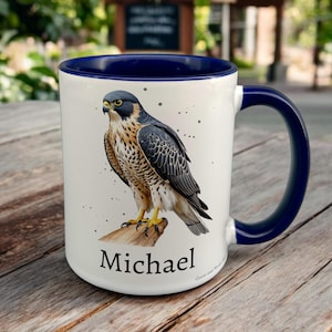Navy & White Ceramic Mug with Falcon Design sat on a table. The Cup has a standard handle. Customisable text is printed below the Falcon design in a black text.