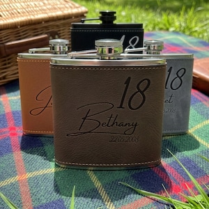 May include: Three metal flasks wrapped in leather-like material in brown, tan, and grey. Each flask features a silver screw-top cap and is engraved with the number 18, a name, and a date. The brown flask is engraved with 18, Bethany, and 22.05.2008.