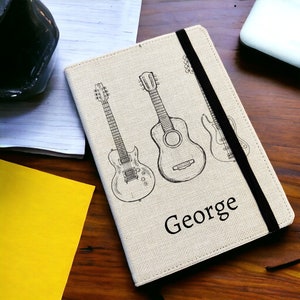 May include: A beige notebook cover with a black elastic closure. The cover features a black and white line drawing of three guitars and the name "George" printed in black.