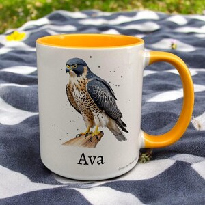 Yellow & White Ceramic Mug with Falcon Design sat on a table. The Cup has a standard handle. Customisable text is printed below the Falcon design in a black text.