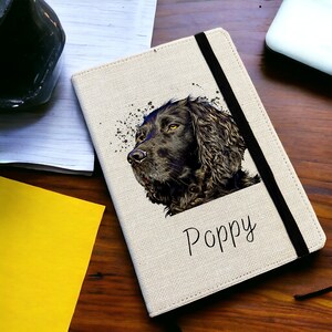 May include: A beige linen notebook with a black elastic closure. The cover features a black and white illustration of a dog with the name "Poppy" printed below.