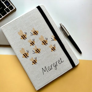 May include: A white canvas notebook with a black elastic closure and a black pen. The notebook features a design of yellow and brown bees and the name "Margret" written in black cursive.