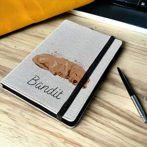 May include: A beige linen notebook cover with a black elastic closure. The cover features a brown dog illustration and the name "Bandit" printed in black.