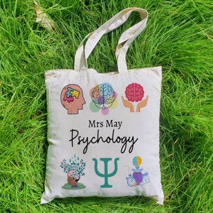 Puede incluir: Bolsa de tela blanca con un dise&ntilde;o colorido que presenta un cerebro, una pieza de rompecabezas, un &aacute;rbol, un s&iacute;mbolo de psicolog&iacute;a y un reloj. El texto "Mrs May Psychology" est&aacute; impreso en la bolsa.