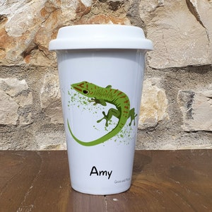 May include: White travel mug featuring a green gecko with red stripes. The mug is personalised with the name "Amy".