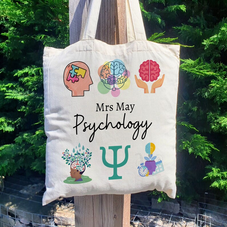 Puede incluir: Una bolsa de lona blanca con un tema de psicolog&iacute;a. Presenta un cerebro, una pieza de rompecabezas, una mano sosteniendo un coraz&oacute;n, un &aacute;rbol con un cerebro, una letra griega psi y una planta en maceta con una bombilla. El texto "Mrs. May Psychology" est&aacute; impreso en la bolsa.