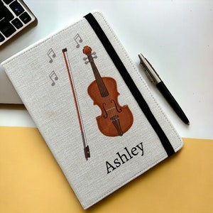 May include: A white canvas notebook with a black elastic closure. The cover features a brown violin with a bow and musical notes. The name "Ashley" is printed in black below the violin.