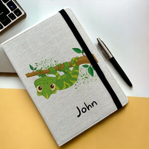 May include: A white canvas notebook with a green chameleon hanging from a branch. The notebook has a black elastic closure and the name "John" is printed on the cover.