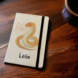 May include: A beige notebook featuring a watercolour illustration of a yellow and orange snake. The name "Leila" is printed in black at the bottom. A black elastic band secures the notebook. The notebook is on a wooden surface.