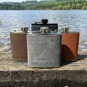 May include: Four metal flasks wrapped in leather-like material in grey, black, and two shades of brown. Each flask features an engraved world map, a small aeroplane icon, and personalised text including names like Emily, Lilly, and Isaac with Gap Year 2027.
