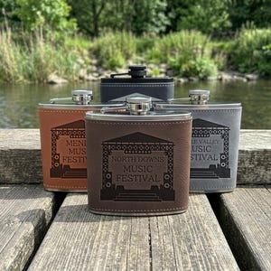 May include: Four stainless steel flasks with leather-like wraps in brown, tan, grey, and black. Each flask features a laser-engraved graphic of a concert stage and the text North Downs Music Festival, Meni Music Festival, or Le Valley Music Festival.