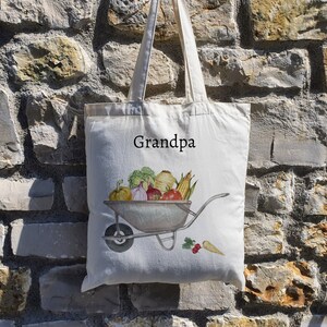 May include: A white canvas tote bag with the word "Grandpa" printed on the front. The bag features a watercolour illustration of a wheelbarrow filled with various vegetables.
