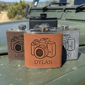 May include: Four metal flasks wrapped in leather-like material in black, brown, tan, and grey. Each flask features a laser-engraved camera illustration and a name, including the name Dylan on the tan flask, Lew on the brown, and Mmy on the grey.