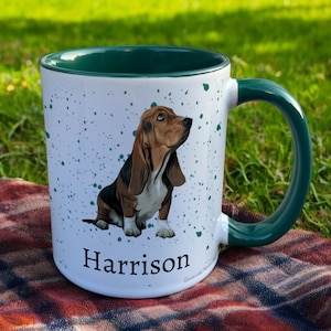 May include: White ceramic mug with a green handle and rim, featuring a Basset Hound illustration and the name "Harrison". The mug has a speckled design and is set against a grassy background.