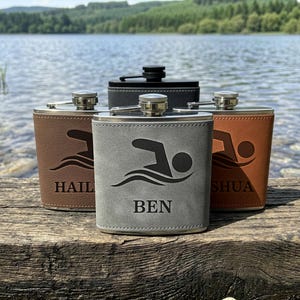 May include: Four metal flasks wrapped in leather-like material, each featuring a laser-engraved swimmer icon and a name. The front flask is grey with the name BEN, flanked by a brown flask labelled HAIL and a tan flask labelled SHUA, with a black flask behind.