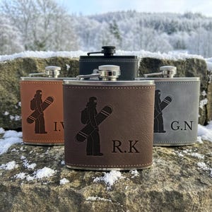 May include: Four metal flasks wrapped in leather-like material in tan, dark brown, grey, and black. Each flask features an engraved graphic of a person carrying a snowboard and personalised initials, including I.V., R.K., and G.N. on the front.