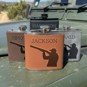 May include: A collection of four stainless steel flasks wrapped in faux leather in brown, tan, and grey colours. Each flask features a laser-engraved silhouette of a person aiming a shotgun, along with a personalised name including Jackson, Harrison, Timothy, and Gerald.