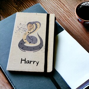 May include: A beige linen notebook cover with a black elastic closure. The cover features a colourful illustration of a snake and the name "Harry" printed in black.