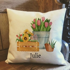 May include: A white cushion featuring a watercolour design of flowers in a wooden crate and a metal bucket. The flowers are pink tulips, yellow sunflowers, and purple flowers. The cushion has the name "Julie" printed on it.