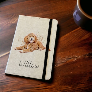 May include: A beige linen notebook with a black elastic closure. The cover features a watercolour illustration of a golden doodle dog with the name "Willow" printed below the image.